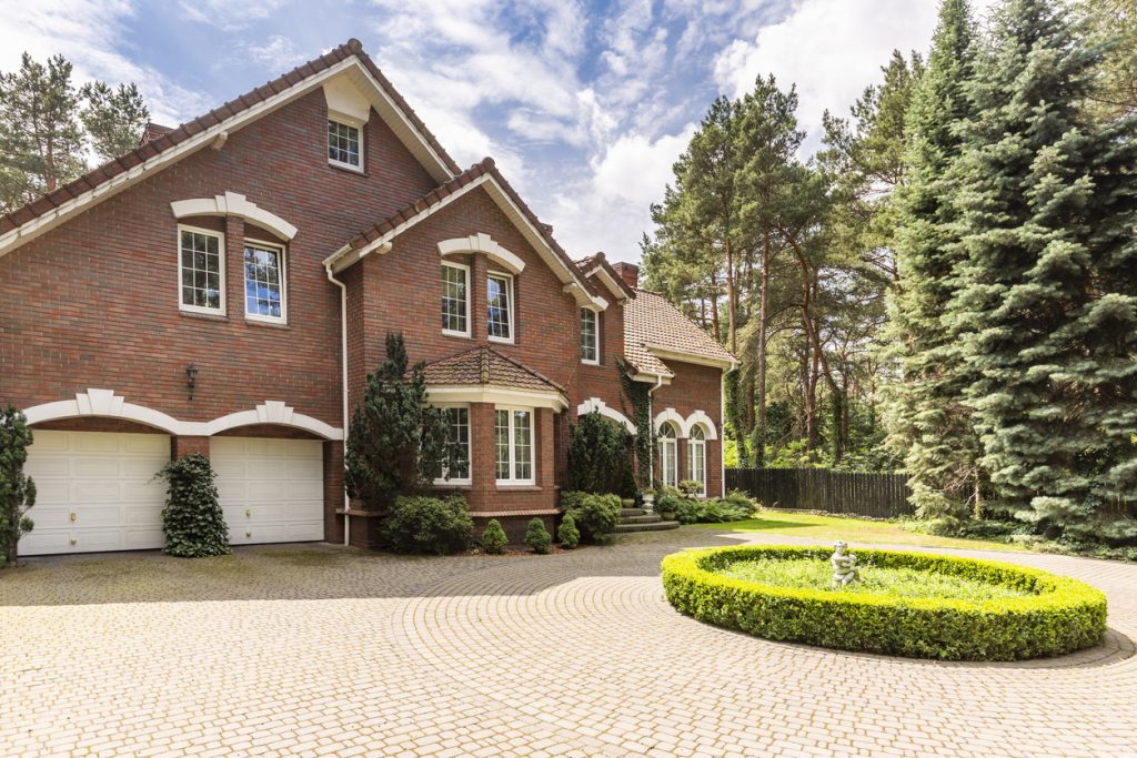 Cobbled circle driveway with green shrubs decoration in front of a traditional red brick English house with steep roof and white windows.